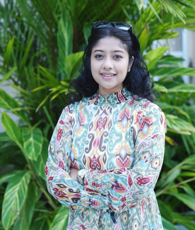 Pinakshi Dutta, founder of House of Tinsel, smiling softly in a patterned ikat dress against green foliage.